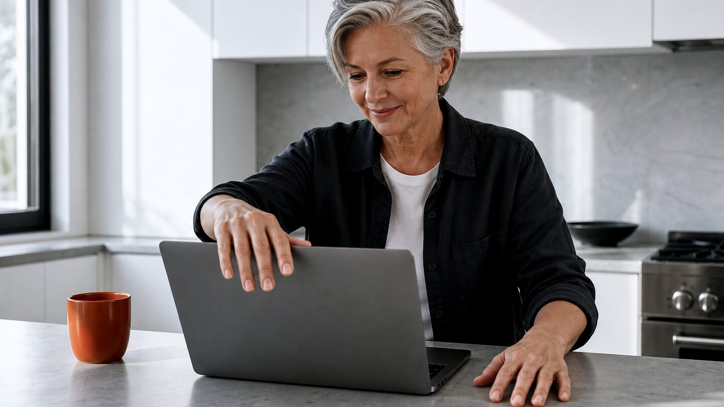 A woman opens a laptop at a kitchen counter, coffee beside her, smiling with anticipation.