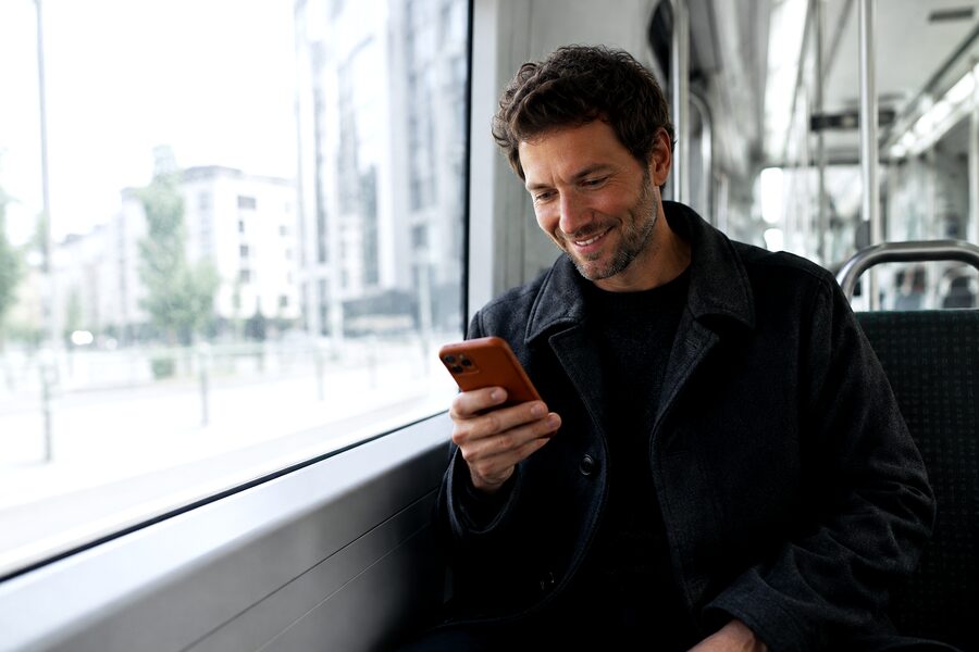 A man rides a tram, phone loosely in hand, smiling at something he just read.